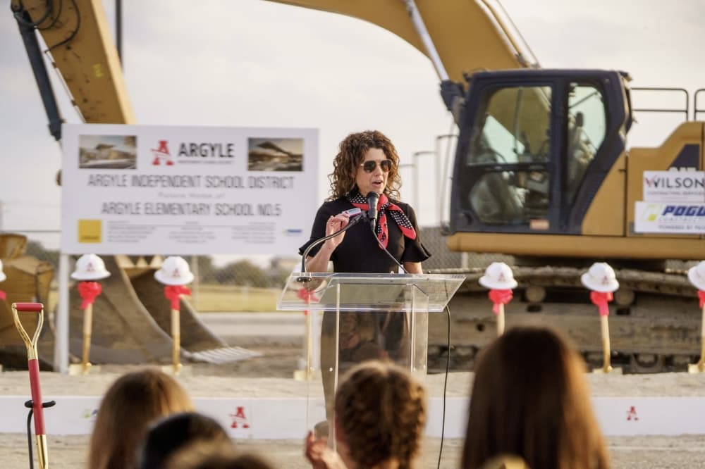 a woman speaks at a podium with construction equipment in the background