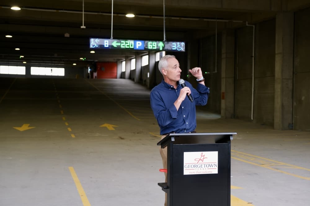 a man holds a microphone and speaks at a podium in a parking garage