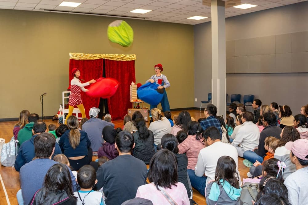 Two people dressed as clowns entertaining an audience sitting on the floor. 