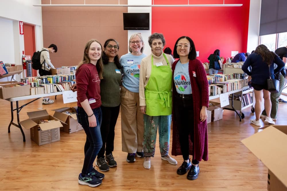 Five females stand in front of tables filled with books. 