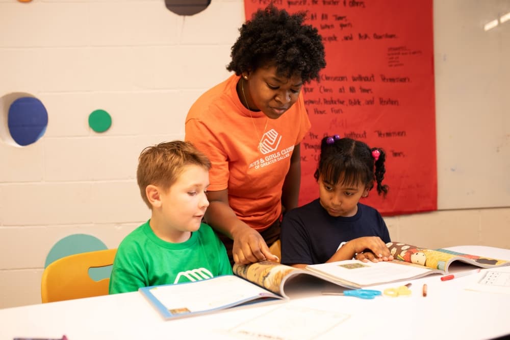 Female adult pointing to a page of a book situated between two children seated at a table. 