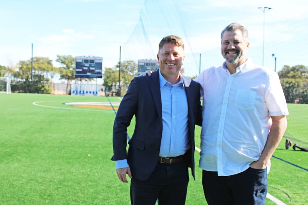 Two males standing side by side next to an outdoor soccer field. 