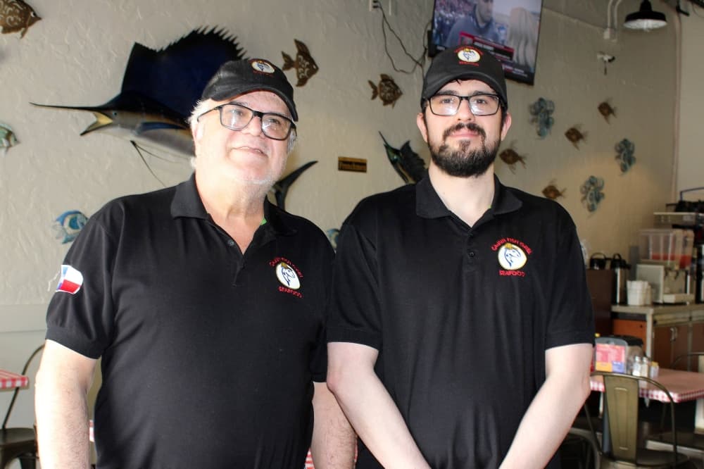 Two males standing side-by-side wearing black shirts featuring Cajun Fish Grill branding. 