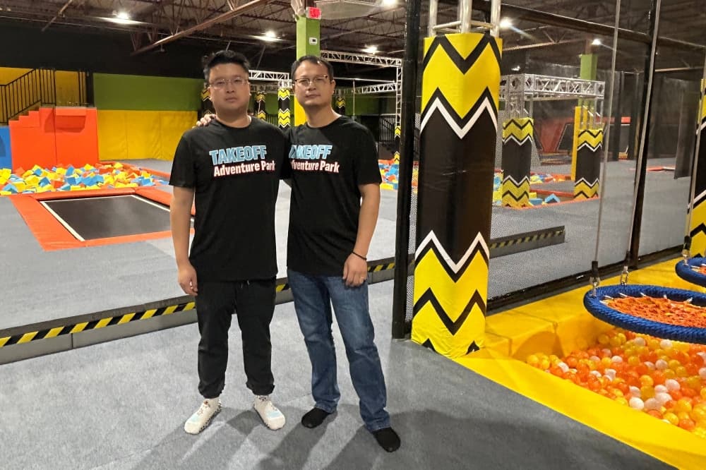 Two males wearing Takeoff Adventure Park t-shirts, standing side-by-side in an indoor playground. 