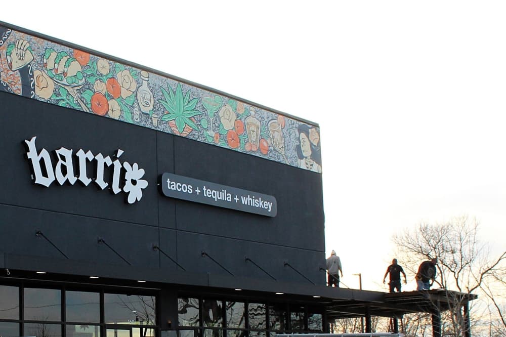 Exterior of a business featuring Barrio signage and three silhouetted figures on top of a overhang attached to the building. 