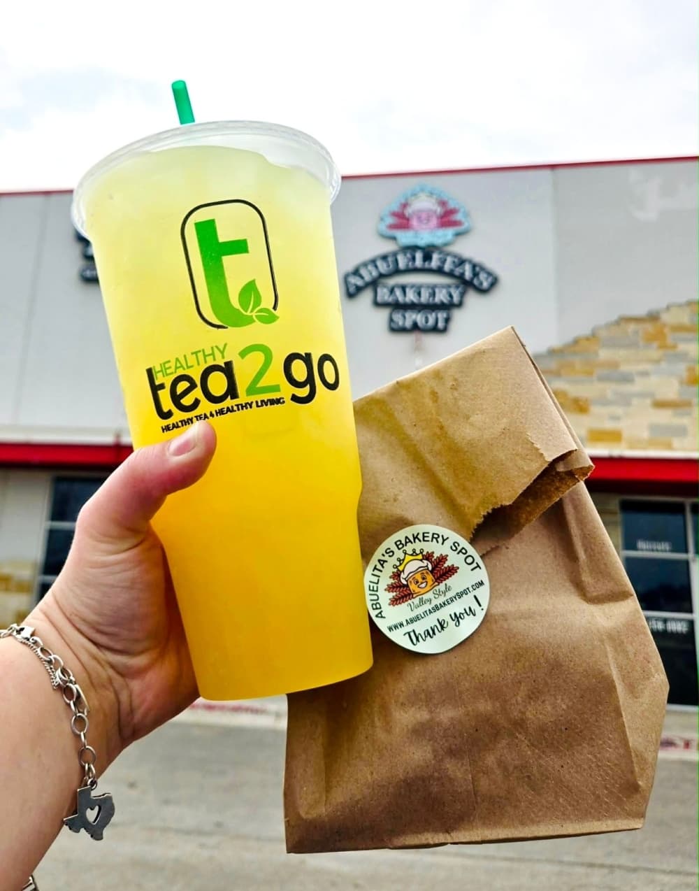 Someone holds up a large yellow Tea2Go TeaN’ergy drink and a brown bag with a white Abuelita’s Bakery Spot sticker.