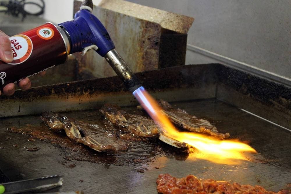 Hand-held flame device being used to torch meat on a griddle.