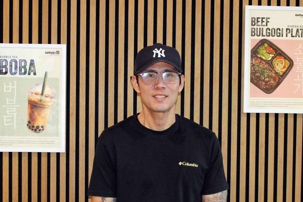 Male standing in front of a brown and black striped wall featuring Korean cuisine posters.