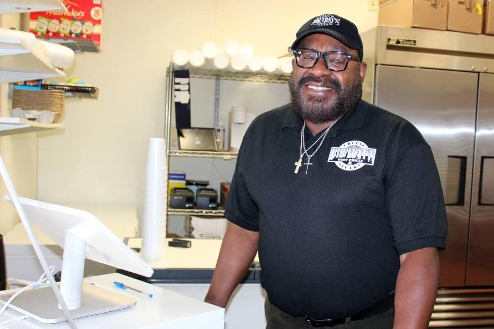 Male wearing a black short-sleeved polo shirt featuring Big Tony's West Philly Cheesesteak branding stands next to a point of sale register. 