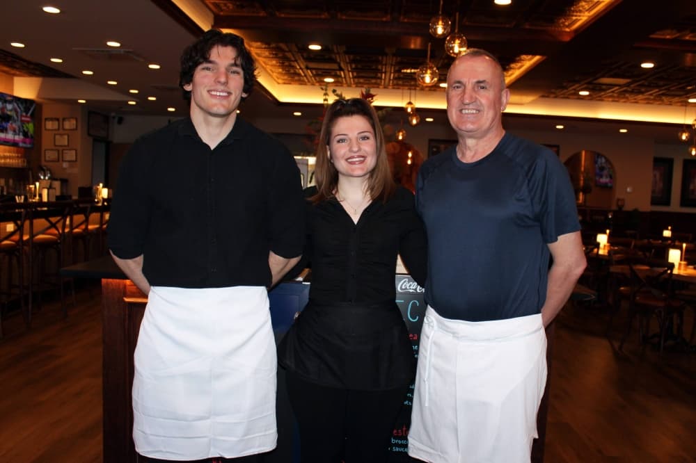Two males standing on either side of a female in a restaurant. All three people are dressed in black and the two men are wearing white waist aprons. 