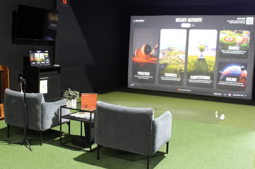 Golf simulator with two chairs facing it and green astroturf floor covering. 