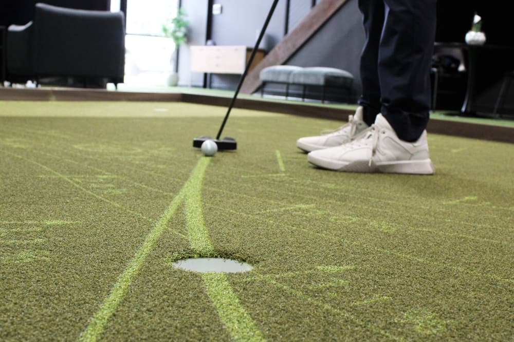 Person's feet and the bottom of a golf club next to a golf ball on a putting green. 