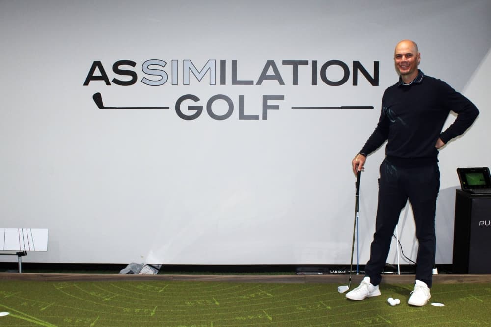 Male holding a golf putter standing on a practice putting green in front of a wall featuring Assimilation Golf signage. 