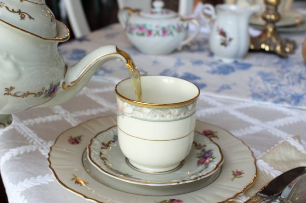 China tea pot pouring tea into a china tea cup sitting on a tea saucer atop a saucer all of which are set on a cloth white table cloth on a table. 