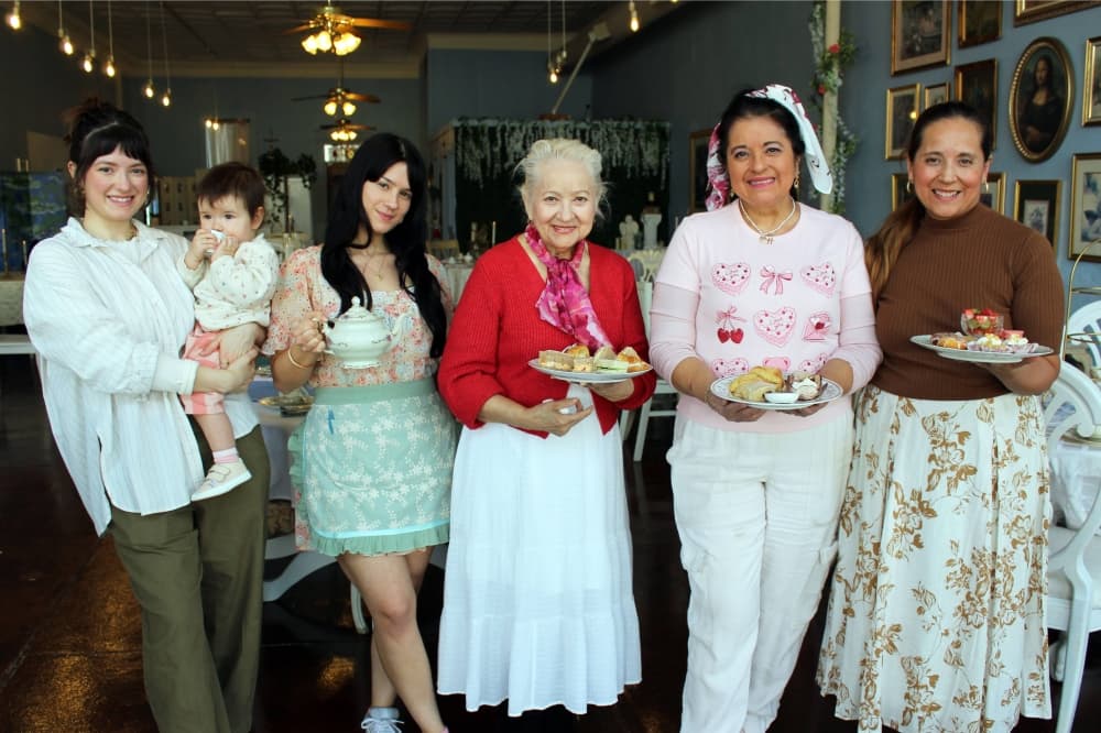 Five women, one of which is holding a baby who is holding a teacup, stand side by side in a restaurant. 