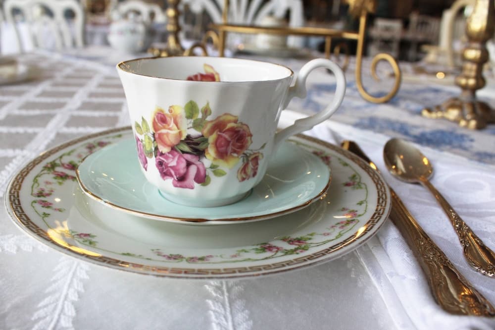 Mismatched tea cup, saucer and plate set on a table featuring a white cloth tablecloth. 