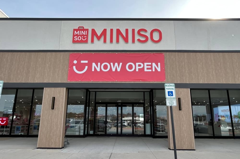 Exterior of a retail center with Miniso permanent signage above a temporary banner featuring Now Open lettering. 