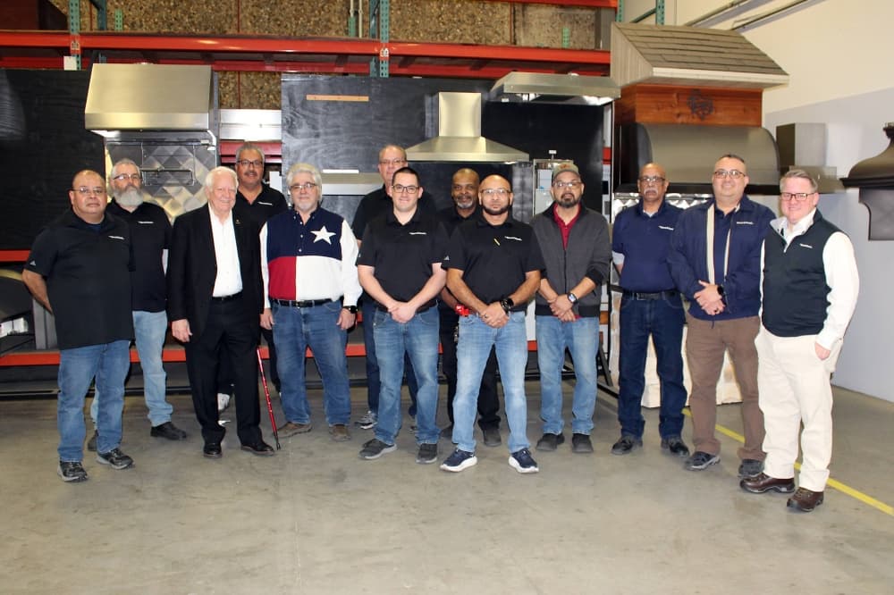 A group of men standing in front in a manufacturing facility in front of stove ventilation canopies. 