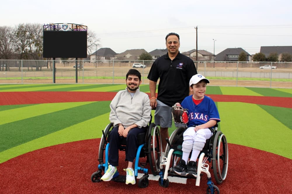 One male standing behind two males sitting in wheelchairs on a baseball field. 