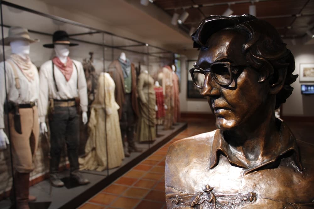 A bronze bust of Larry McMurtry stands in front of cowboy outfits.