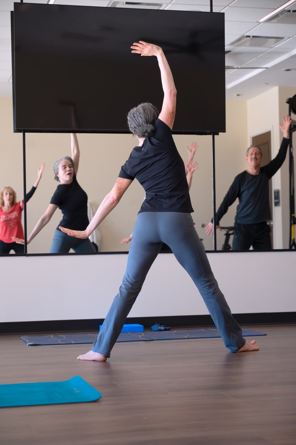 Debra Butts leads students through a flowing movement sequence, her reflection showing the class following along in real time.
