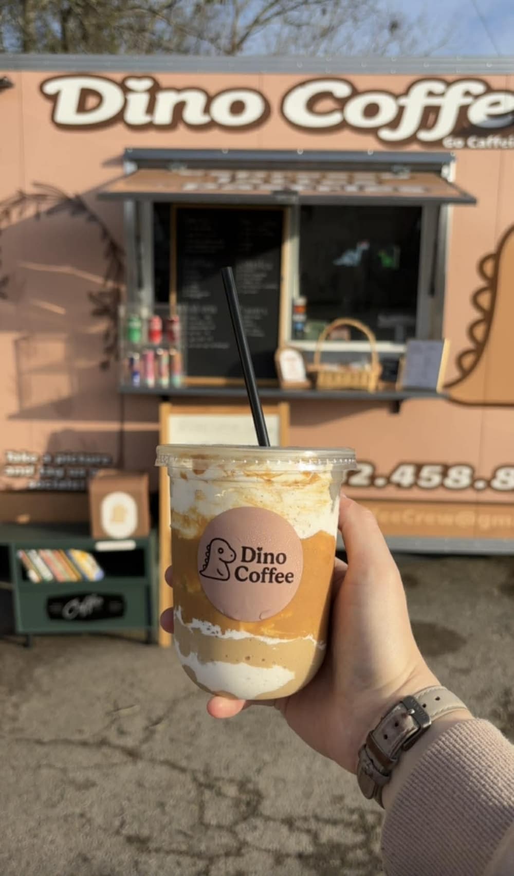Dino Coffee company iced latte—with layers of caramel and whipped cream in a see-through cup—being held in front of the business's trailer