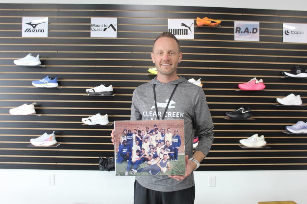 Man standing in front of a wall of running shoes holds a photo of his high school cross-country team