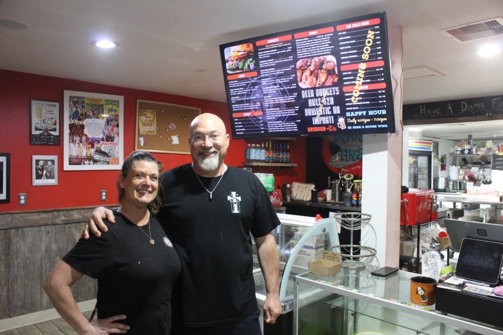 Husband and wife stand in front of their restaurant's menu 