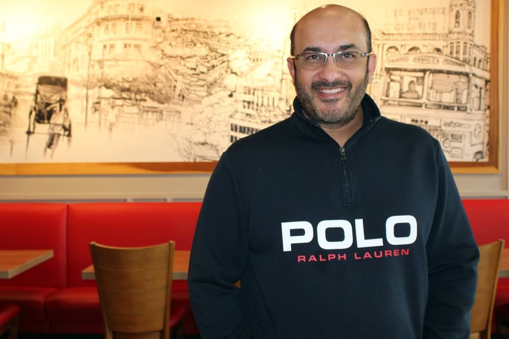 Male standing in a restaurant in front of a mural depicting a city scene line drawing. 