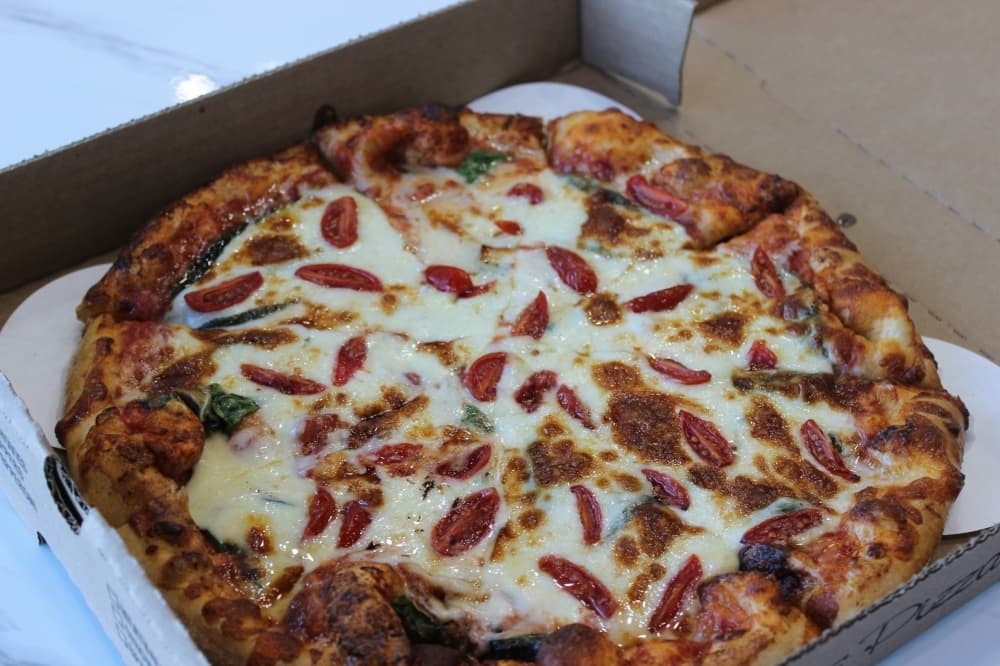 Large pizza in a takeout box topped with mozzarella cheese, grape tomatoes, fresh basil, and herbs on a lightly charred crust.