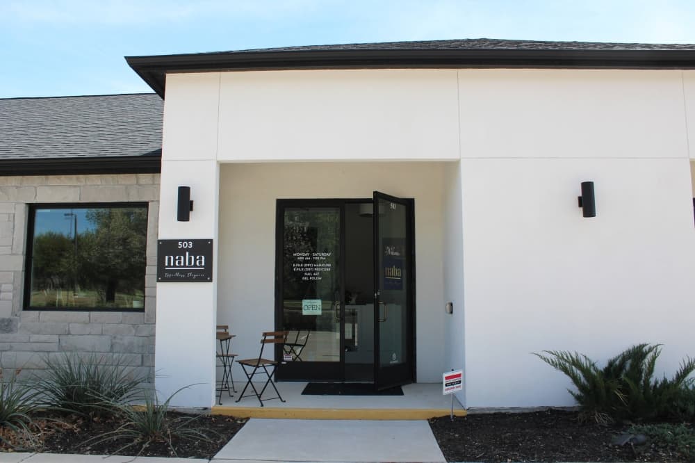 Exterior of white building with black roof and architectural elements featuring a Naba Style Nails sign affixed to the building. 