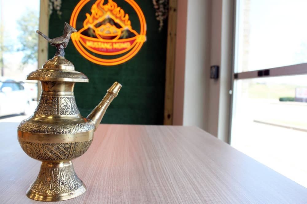 Ornate brass pouring receptacle placed on a table in front of a photo wall featuring Mustang Momo & Grill branding. 