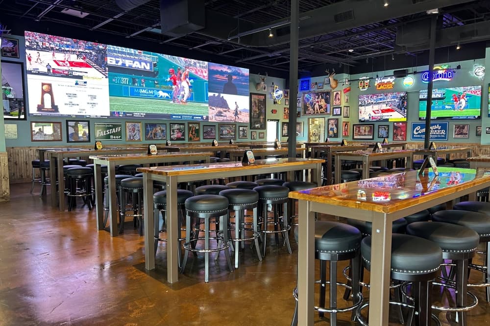 Interior of a restaurant featuring large TV screens and high top tables with barstools. 