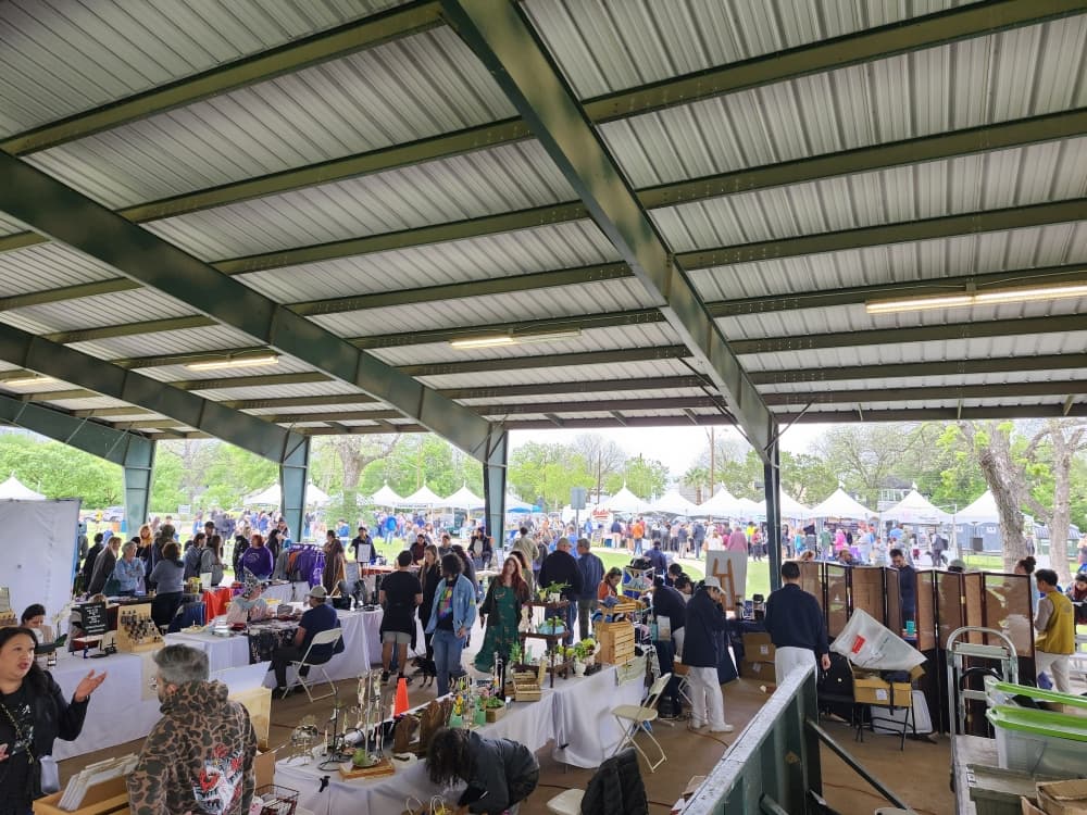 Crowds browse vendor tables inside a large open-air pavilion, with white event tents and green trees visible in the background.