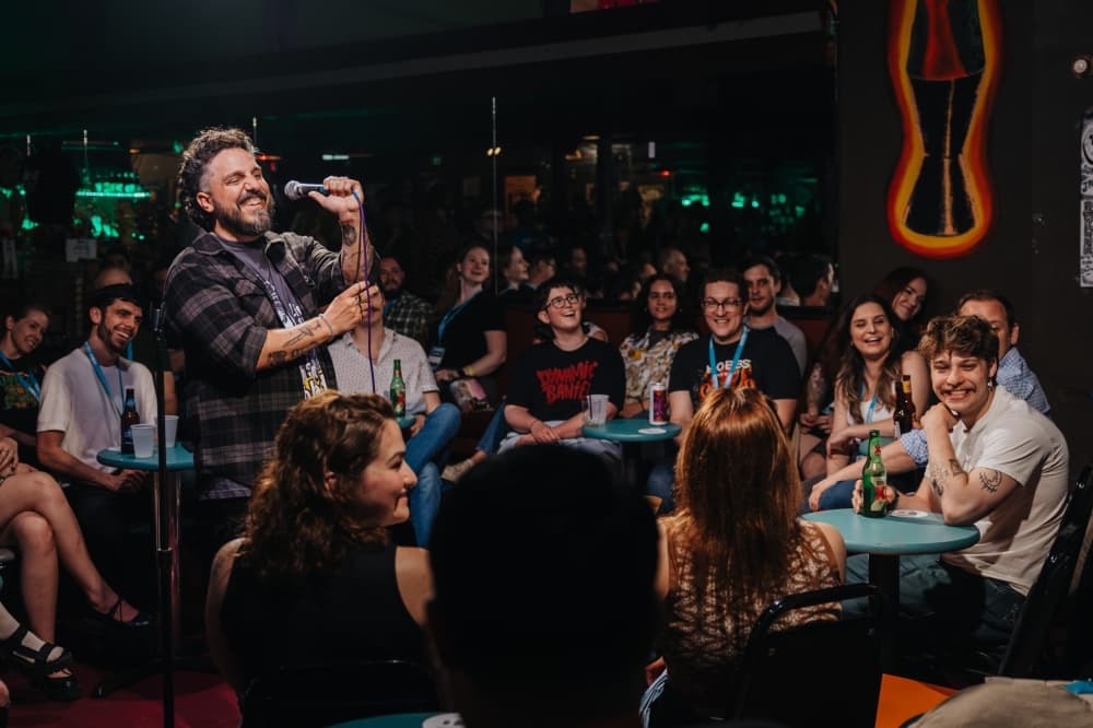 A bearded comedian holds a microphone and smiles at a packed bar audience seated at small round tables.