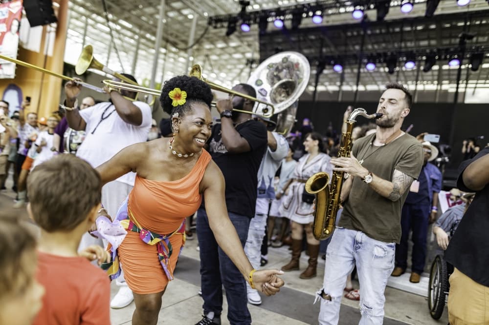 A woman in an orange dress dances alongside a brass band performing in front of a stage at an outdoor music festival.