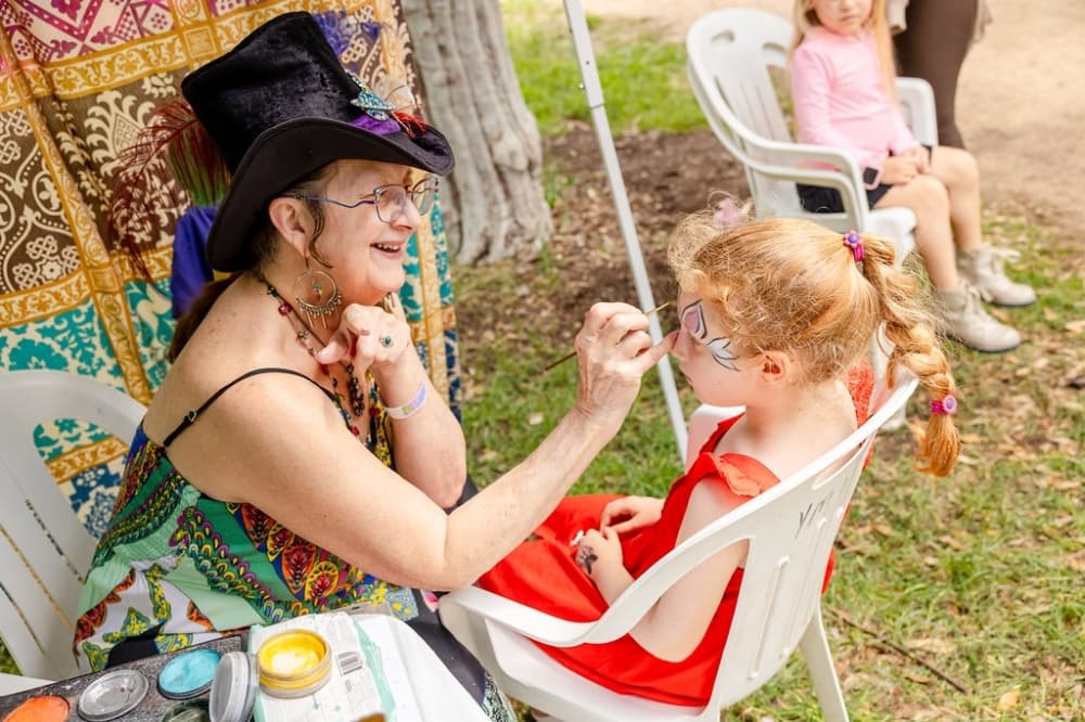 A face painter in a wide-brimmed black hat applies paint to a young girl's face at an outdoor festival booth.