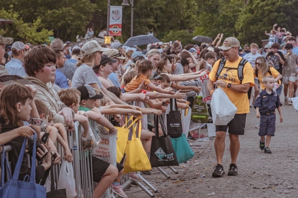 A man in a yellow shirt hands items to children and adults reaching over a barricade along a crowded outdoor parade route.