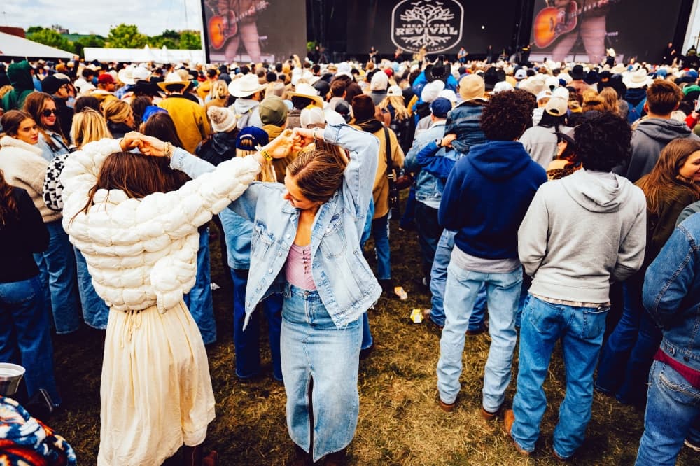 A dense crowd faces an outdoor stage at a country music festival, with several attendees in cowboy hats and denim.