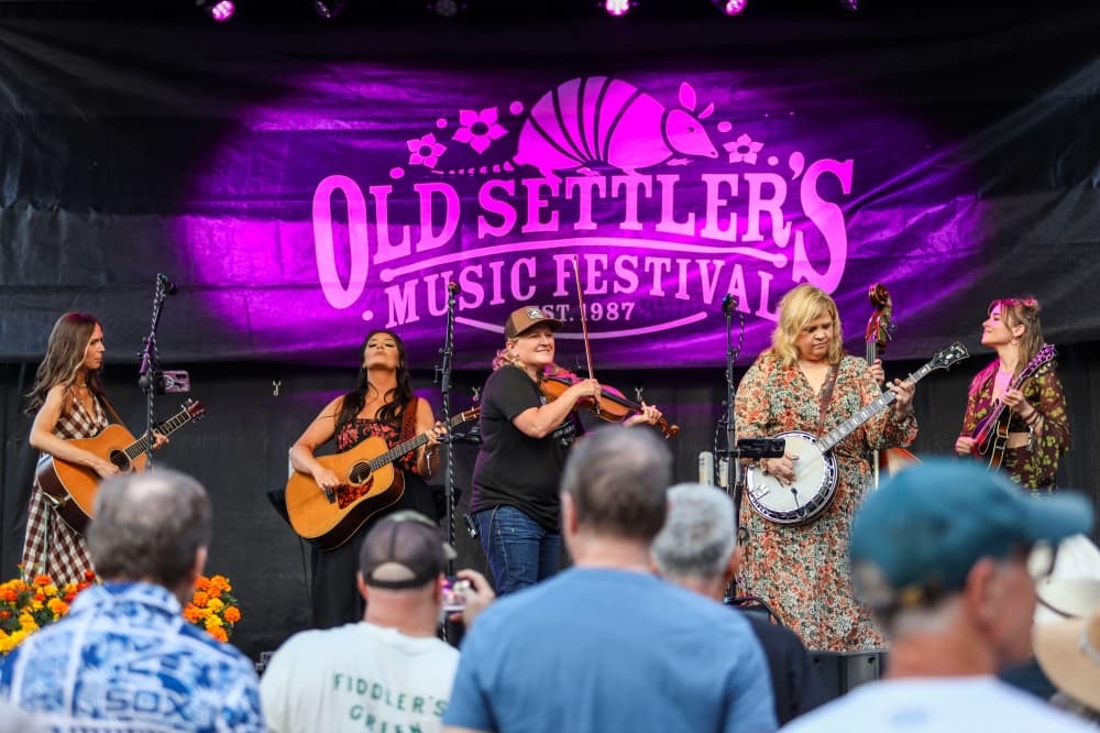 Five women perform on an outdoor stage beneath a large illuminated Old Settler's Music Festival banner, playing acoustic guitar, fiddle, banjo, and upright bass.