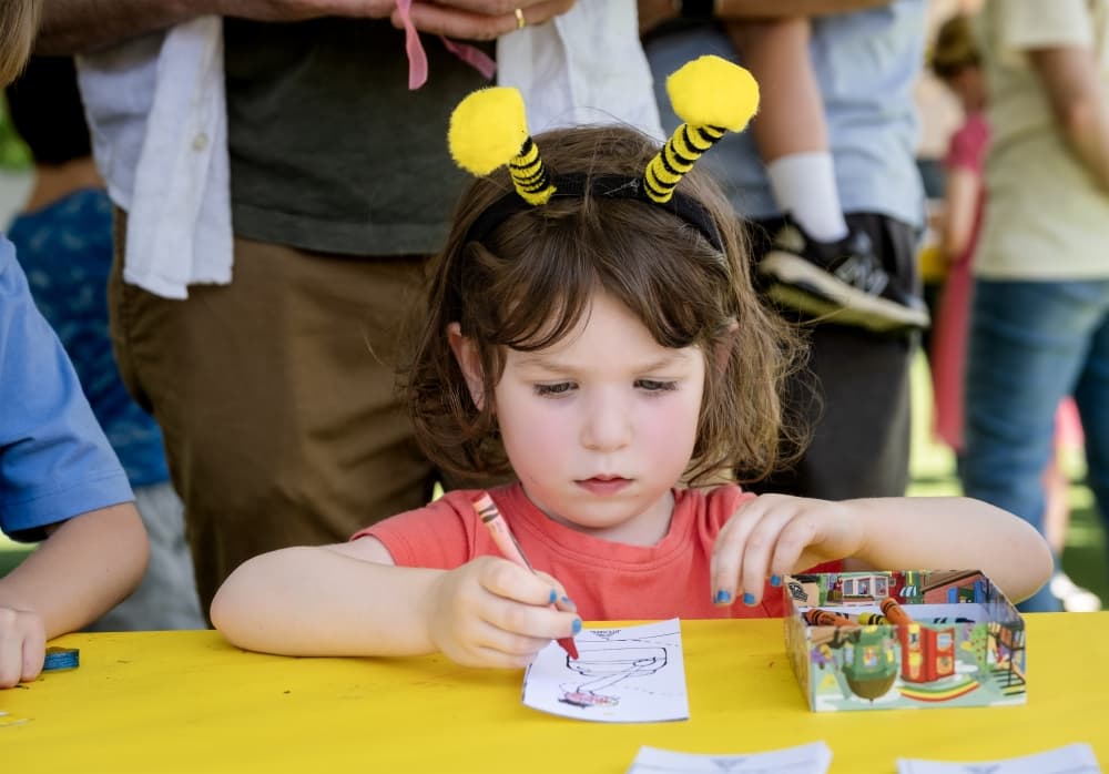 A young child wearing bee antenna headband sits at a yellow table, concentrating on a coloring activity.