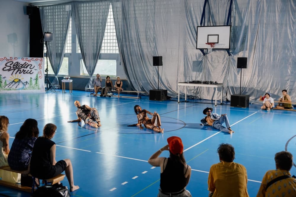 Three dancers lie and recline on a blue gymnasium floor mid-performance, with an audience seated around the edges of the court and a basketball hoop visible overhead.