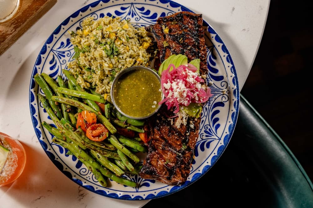 A blue and white plate of skirt steak topped with avocado, pickled red onions and cheese, cilantro line rice, and asparugus.