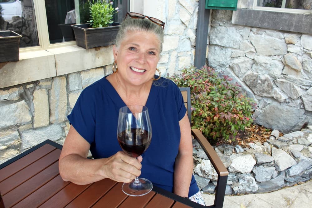 Female holding a class of wine while seated at a wood table in a patio setting. 