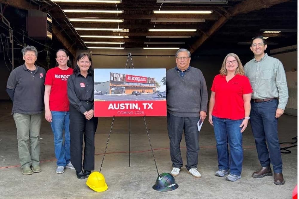 Six people stand with a rendering of the new Half Price Books location: a woman with short hair in a black shirt, a woman in jeans and a red Half Price Books shirt, a woman in a black button up and a red name tag, a man in black and in a grey sweater, a woman with glasses in a red tee shirt and jeans, and a man in a grey button up and glasses. There are two hard hats in front of the sign, located in an empty garage.