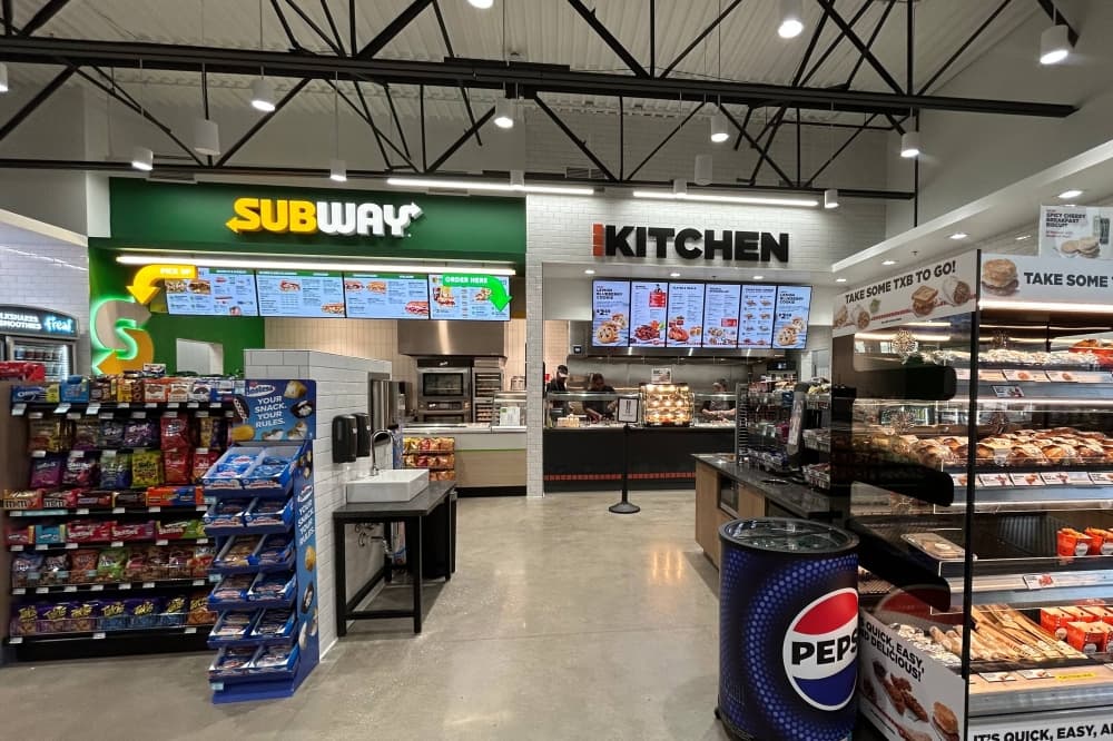 Interior of a convenience store with a Subway counter and another restaurant ordering counter featuring the word Kitchen above the counter.