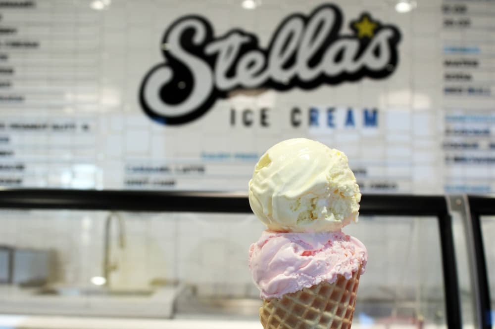 Two scoops of ice cream in a waffle cone in front of a wall featuring Stella's Ice Cream branding. 
