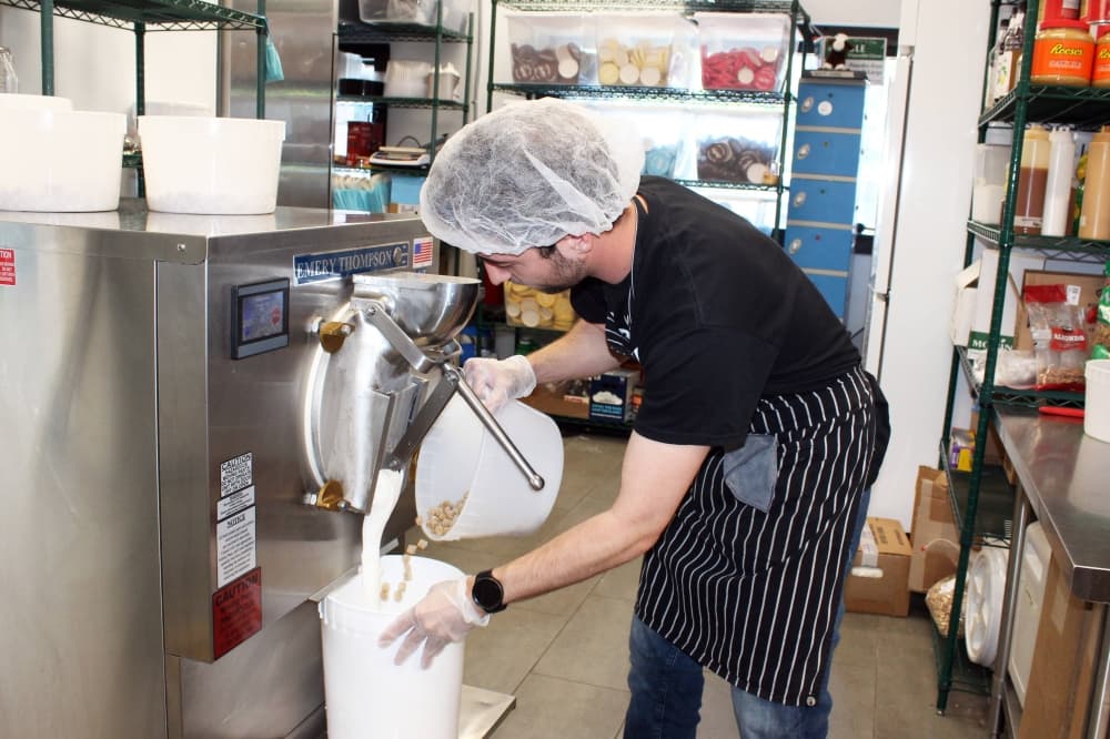 Male wearing a hair net making cookie dough ice cream in a retail space. 