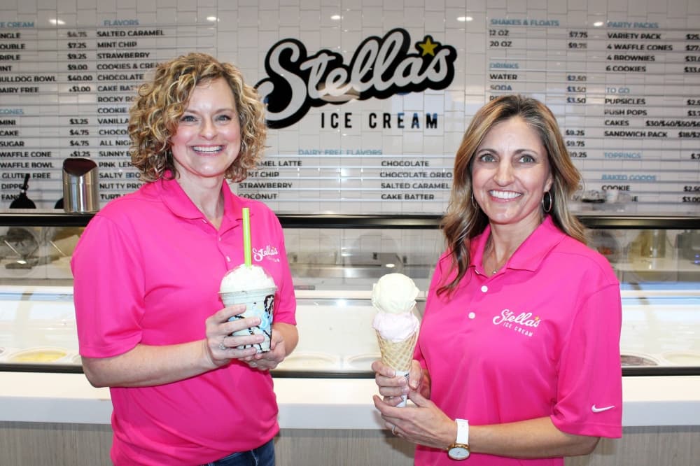 Two females wearing bright pink polos featuring Stella's Ice Cream branding standing next to each other in an ice cream shop while holding scoops of ice cream in a cone and a milkshake.