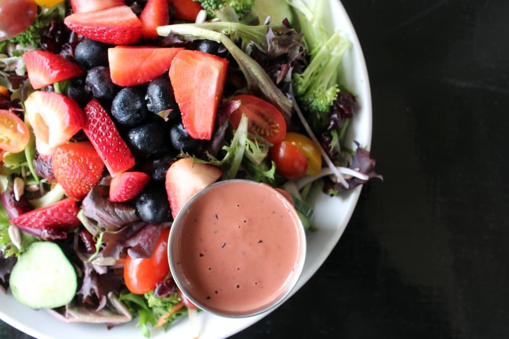 Overview of a salad including strawberries, blueberries, lettuce and container filled with pink salad dressing. 